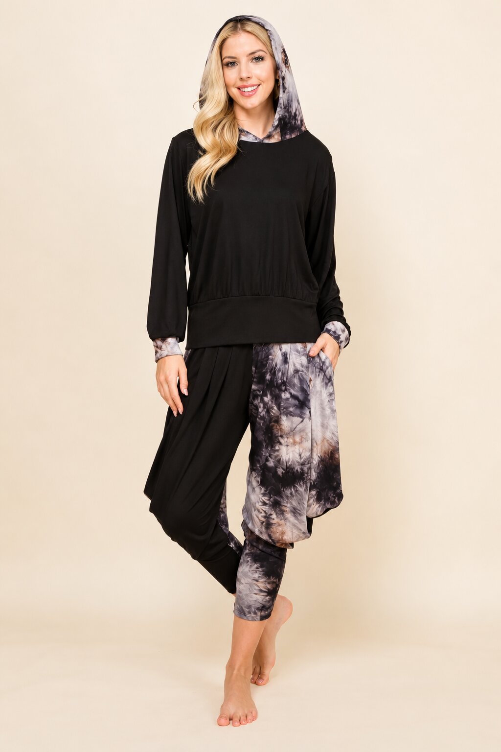 A woman modeling a two-piece lounge set featuring a black long-sleeve top with a matching patterned hood and black jogger pants with gray and black tie-dye patterned leg warmers, against a solid beige background.