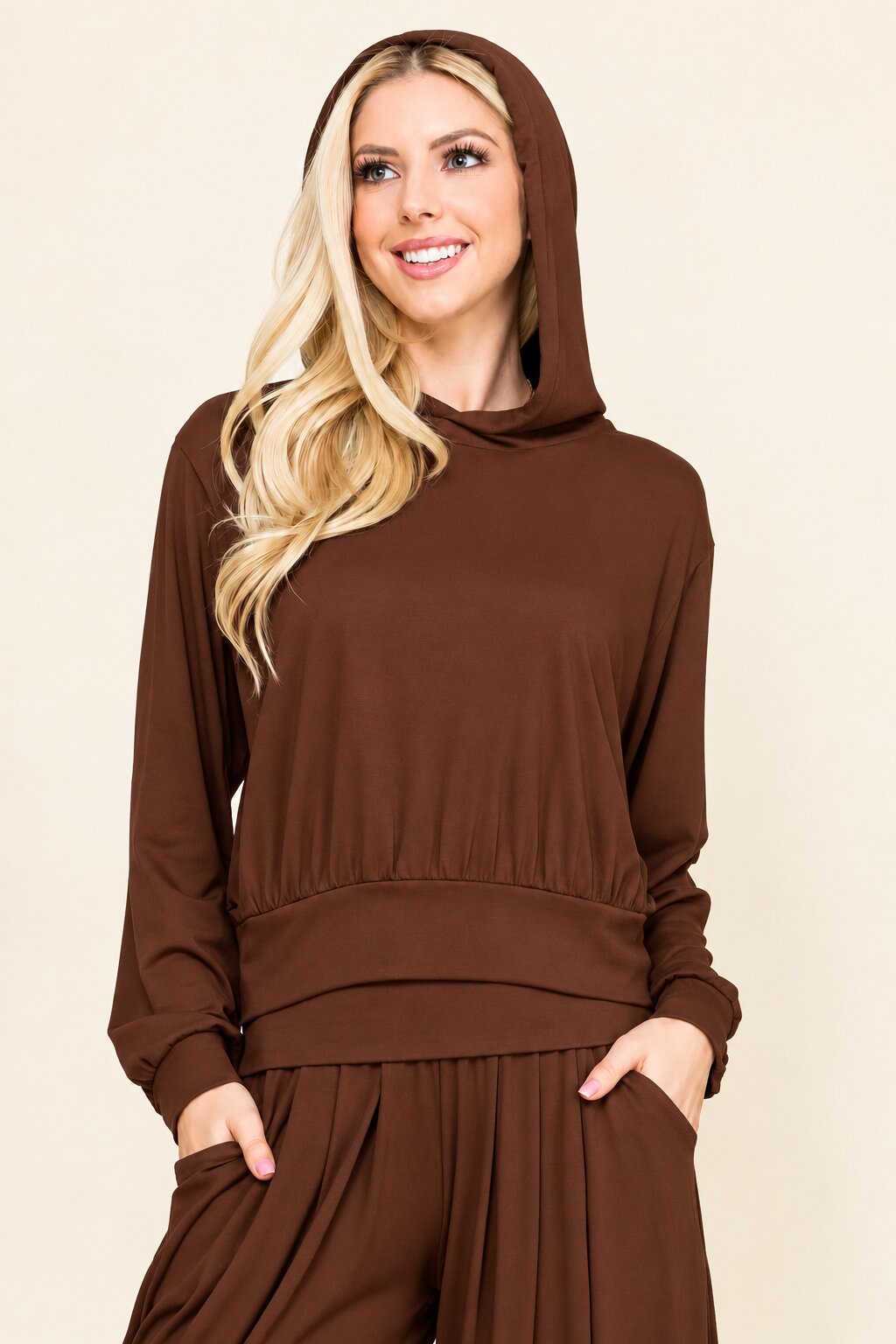 A woman wearing a matching brown hooded sweatshirt and pleated pants set, standing against a solid beige background.