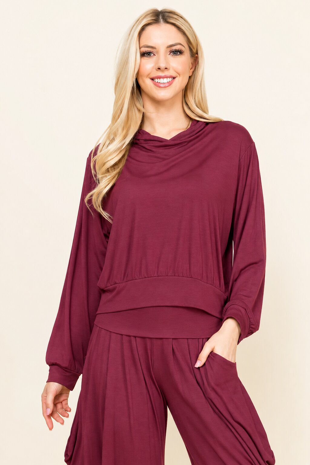 A woman modeling a matching two-piece loungewear set in deep burgundy, featuring a long-sleeve cowl-neck top and coordinating drawstring pants, set against a plain beige background.