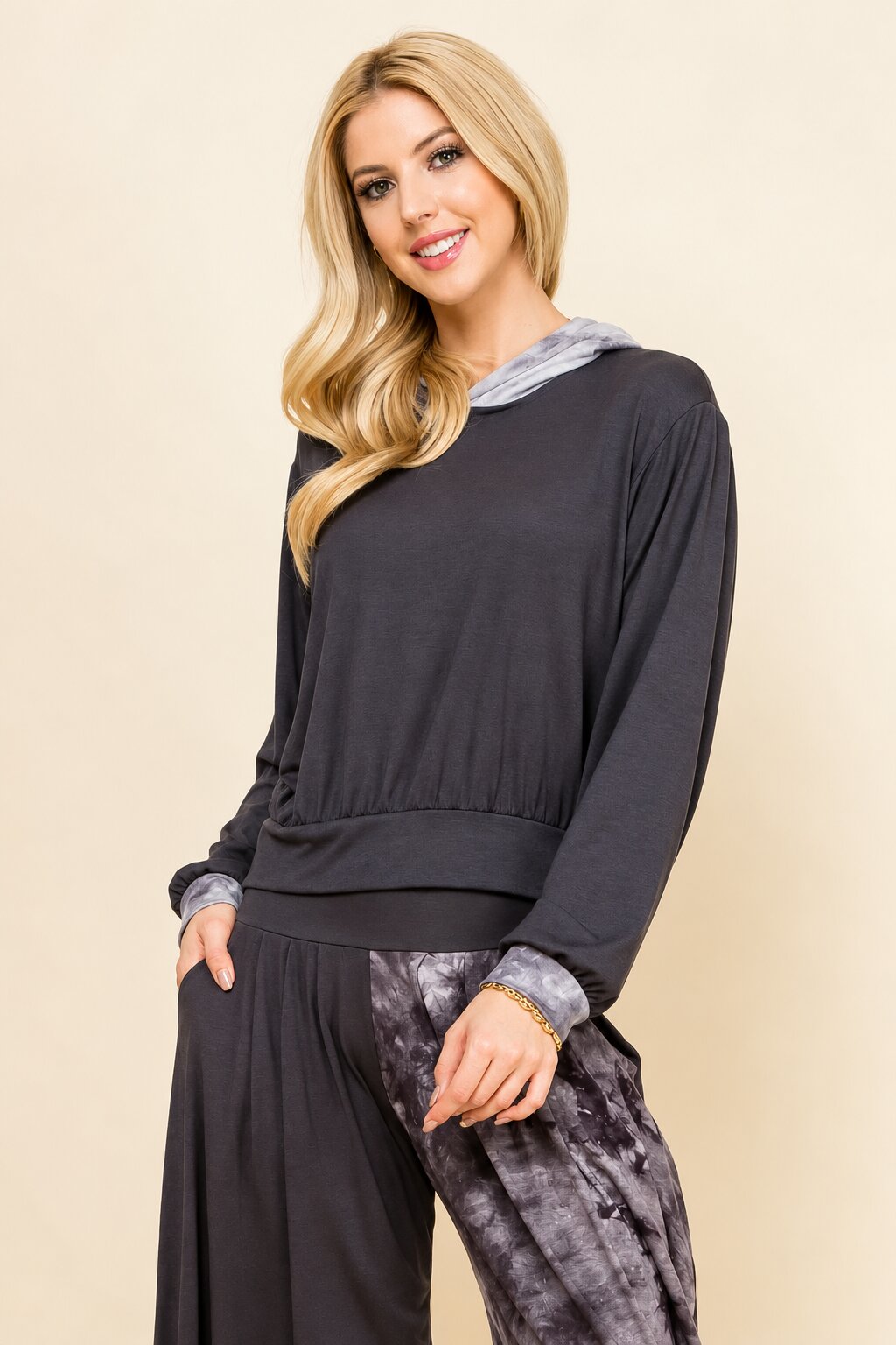 A woman modeling a dark charcoal gray long-sleeve top with a light gray hood and matching dark gray wide-leg pants, set against a solid beige background.