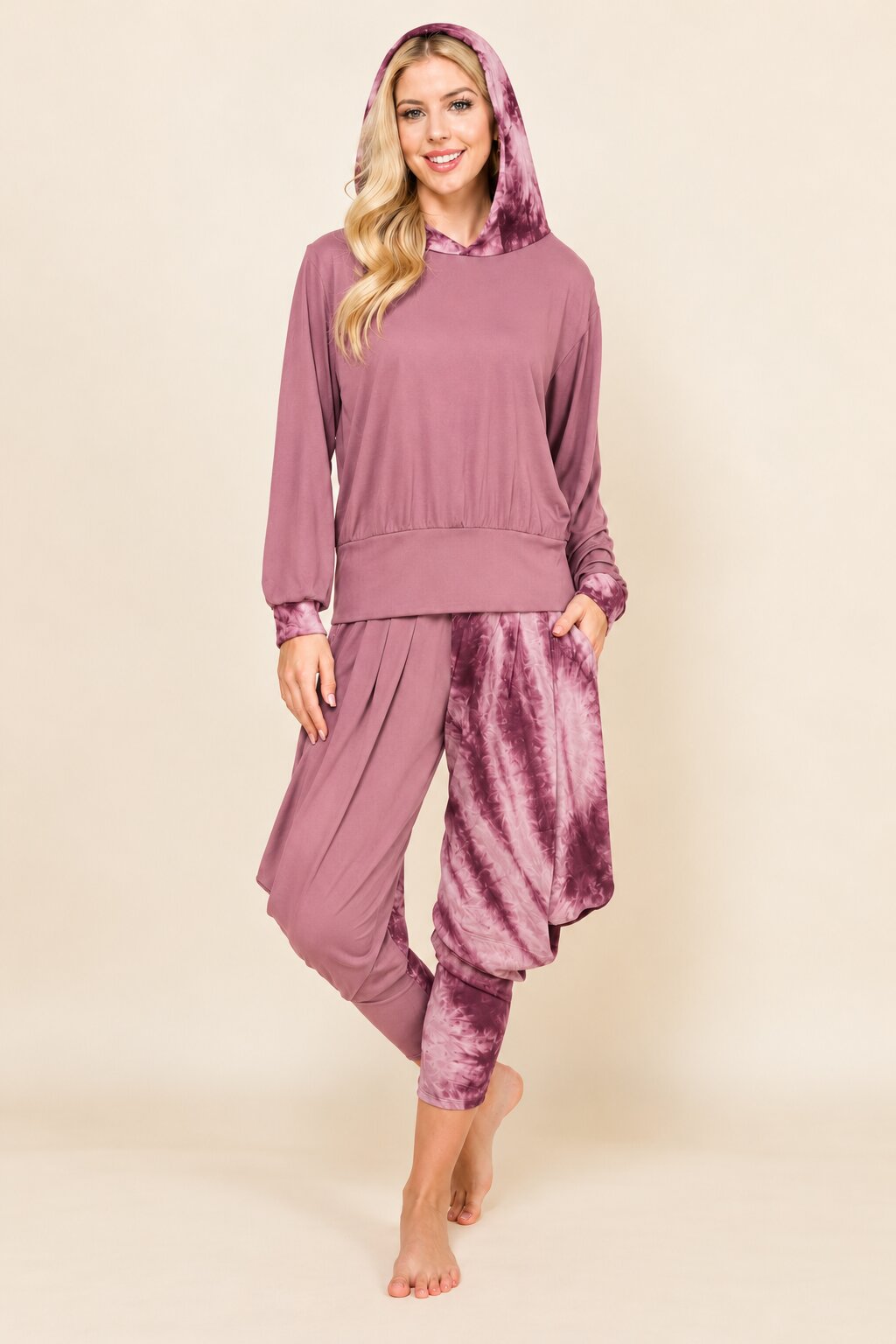 A woman modeling a mauve-colored two-piece loungewear set consisting of a long-sleeve hooded sweatshirt and matching relaxed pants with a velvet-like texture, against a solid beige background.