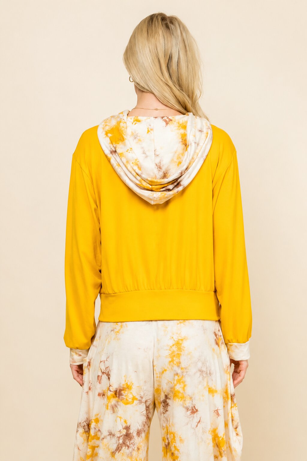 A woman shown from the back wearing a bright yellow hooded sweatshirt with a white and yellow tie-dye patterned hood, paired with matching tie-dye pants, against a solid beige background.