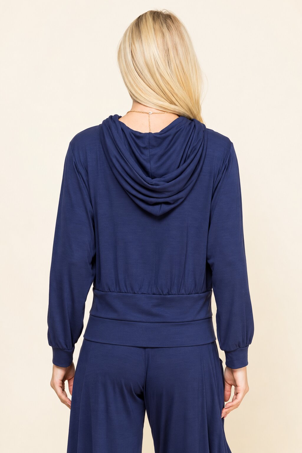 A woman viewed from the back wearing a navy blue draped hooded long-sleeve top and matching pants against a light beige studio background.
