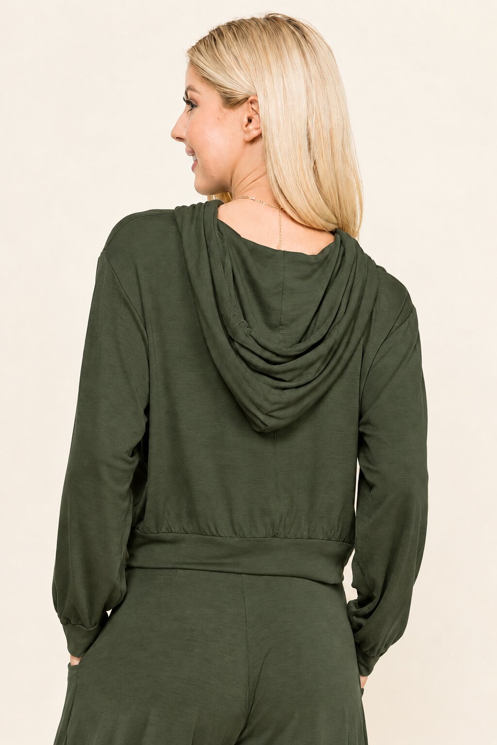 A woman shown from the back wearing an olive green long-sleeve top with a draped hood or collar, paired with matching green bottoms, against a plain light beige studio background.