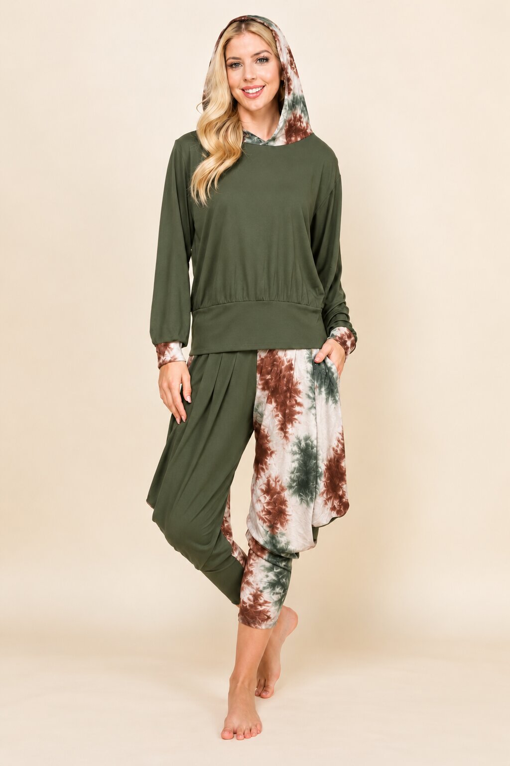 A woman modeling a two-piece lounge set consisting of an olive green long-sleeve top with a matching patterned hood and distressed olive green pants featuring a brown, green, and white tie-dye side panel, set against a solid beige background.