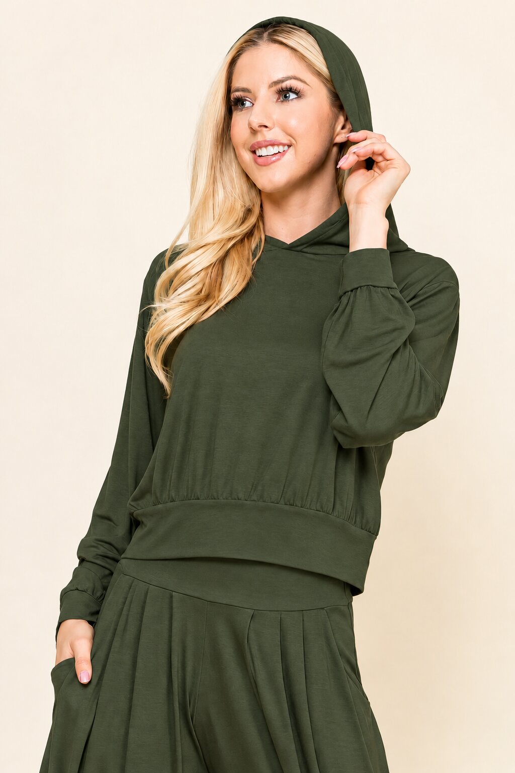 A woman modeling an olive green two-piece loungewear set consisting of a long-sleeve hooded sweatshirt and matching flared pants, set against a plain beige background.