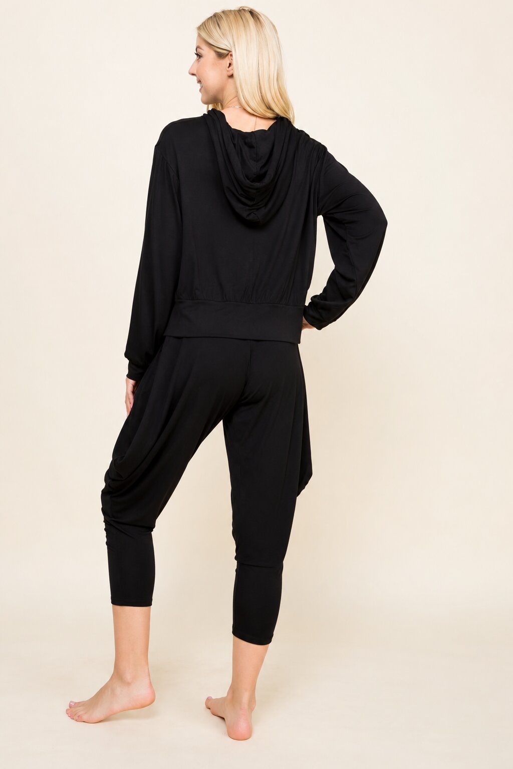 A woman modeling a black two-piece loungewear set, featuring a draped long-sleeve top and matching cropped pants, shown from a rear three-quarter angle against a solid light beige background.