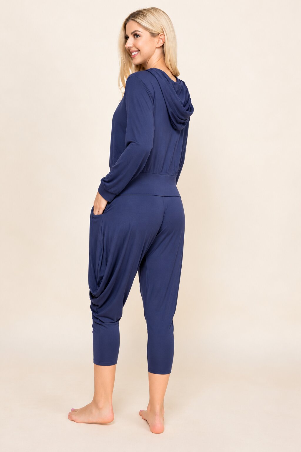 A woman modeling a navy blue hooded jumpsuit with long sleeves, side pockets, and draped capri-length legs, shown from a side-back angle against a plain beige studio background.