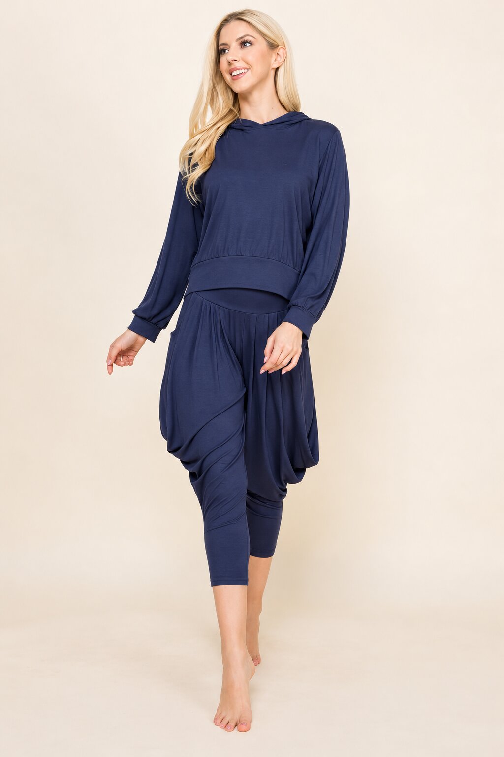 A woman modeling a matching navy blue loungewear set consisting of a long-sleeve cropped hoodie and draped capri pants, set against a solid beige studio background.
