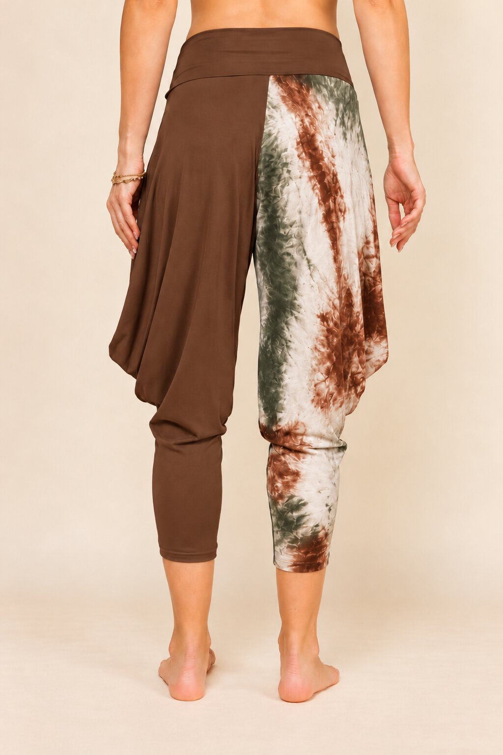 Rear view of a person wearing harem-style cropped pants with a solid brown left side and a brown, green, and off-white tie-dye pattern on the right side, against a plain beige studio background.