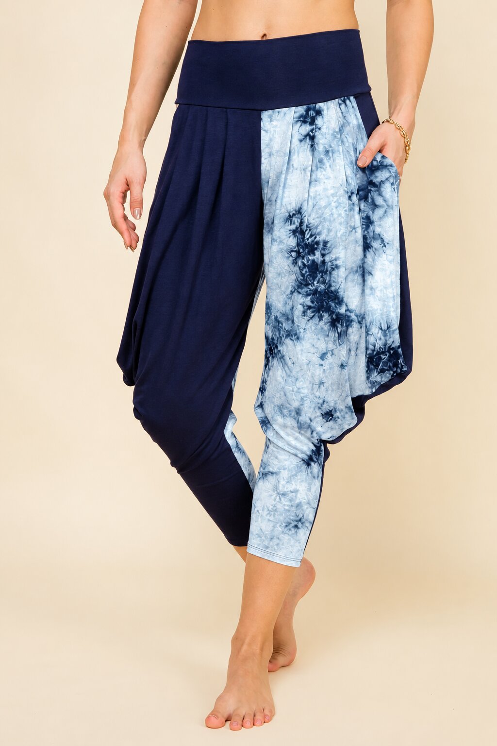 A person modeling loose harem-style cropped pants with a solid navy left leg and a blue-and-white tie-dye right leg, shown against a plain beige studio background.