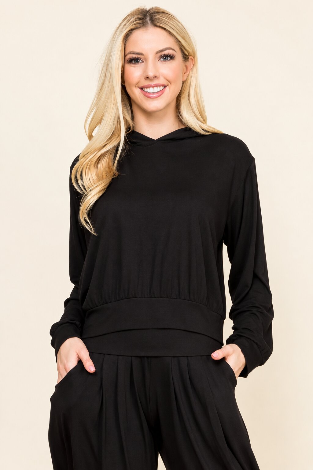 A smiling blonde woman wearing a black long-sleeve hooded sweatshirt and black pants, standing against a plain beige studio background.