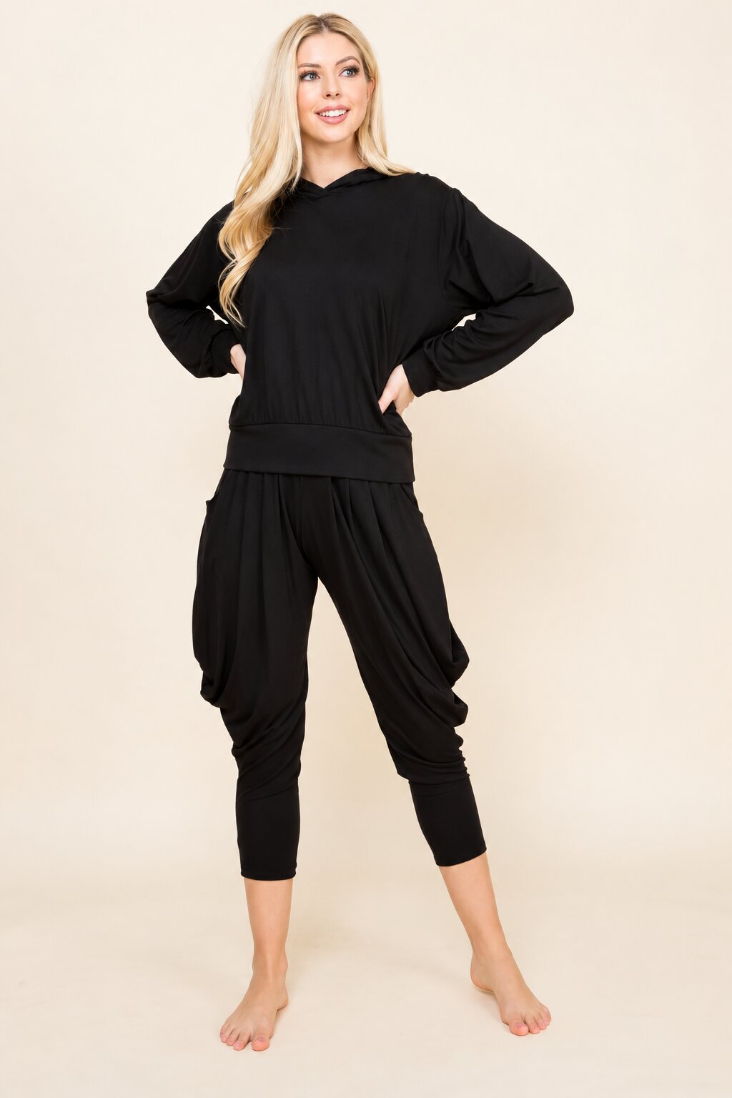 A woman modeling a black two-piece loungewear set consisting of a long-sleeve cropped hoodie and matching draped capri pants, standing against a solid beige studio background.