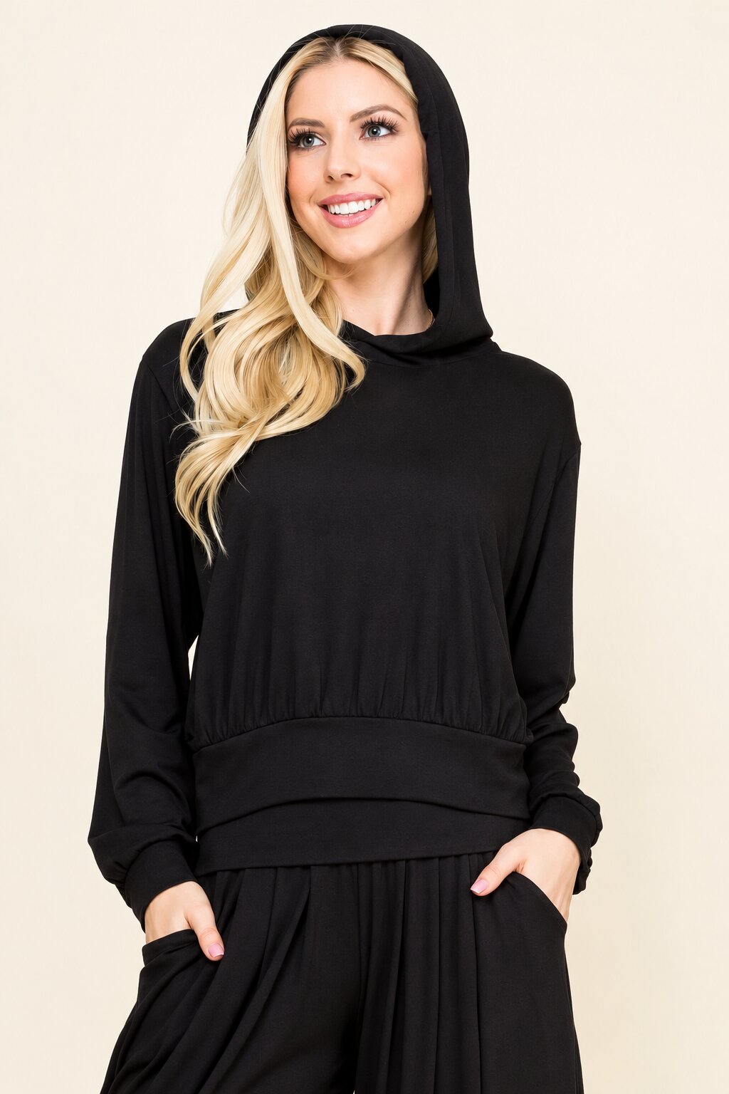 A woman with blonde hair wearing a black long-sleeve hooded top and matching black pants, standing against a plain beige studio background.