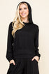A woman with blonde hair wearing a black long-sleeve hooded top and matching black pants, standing against a plain beige studio background.