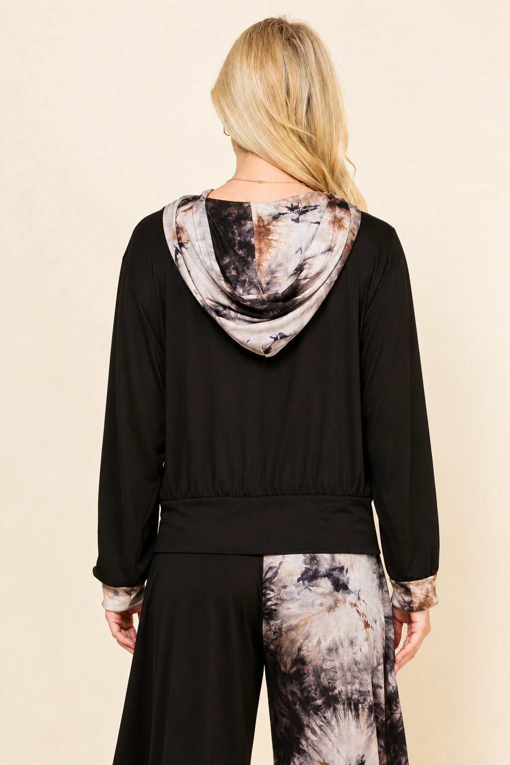A woman shown from the back wearing a black long-sleeve top with a hood featuring a gray, white, and brown tie-dye pattern, paired with matching patterned pants, against a solid beige background.