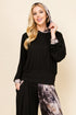 A woman modeling a black long-sleeve top with patterned cuffs and a matching headscarf, paired with black pants featuring a gray and brown abstract print, against a solid beige background.