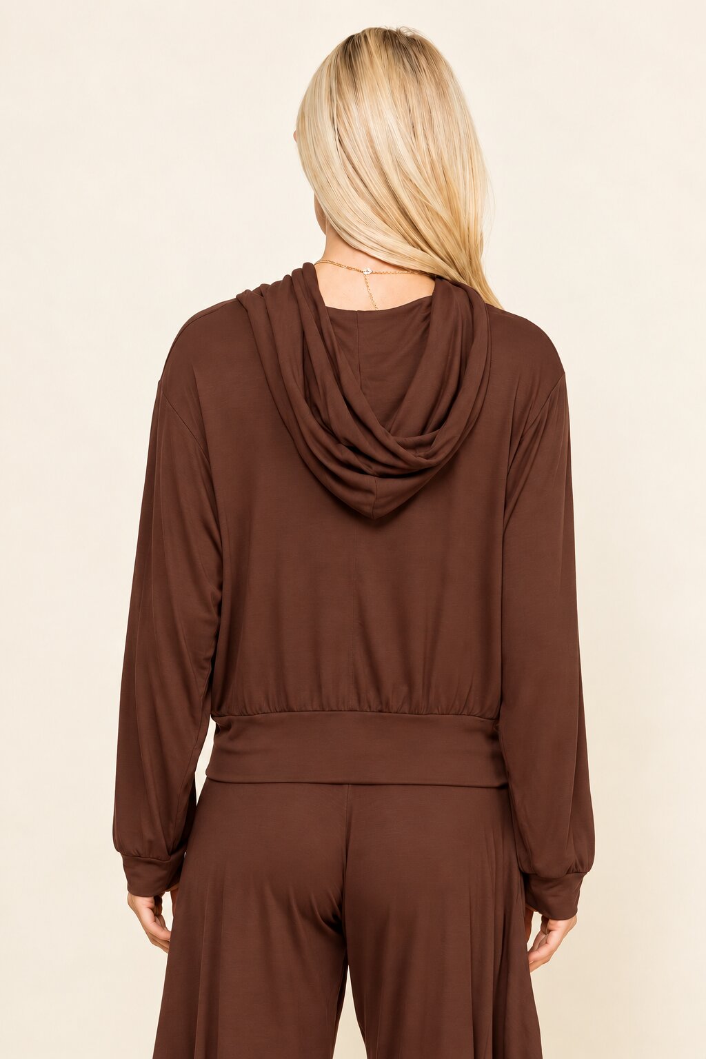 A woman viewed from the back wearing a brown long-sleeve top with a draped cowl neckline and matching brown bottoms, against a plain light beige studio background.