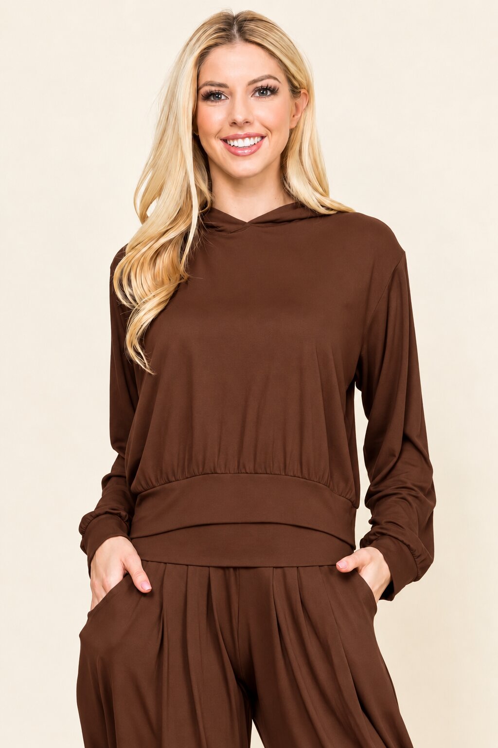 A smiling blonde woman wearing a matching brown long-sleeve hooded top and brown wide-leg pants, standing against a plain beige studio background.