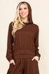 A woman wearing a matching brown hooded sweatshirt and pleated pants set, standing against a solid beige background.