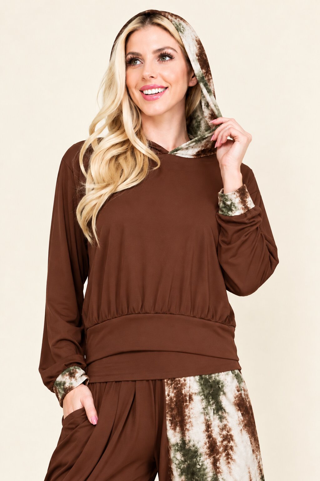 A woman wearing a brown long-sleeve top with a matching brown and green patterned headscarf and patterned pants, posed against a solid beige background.