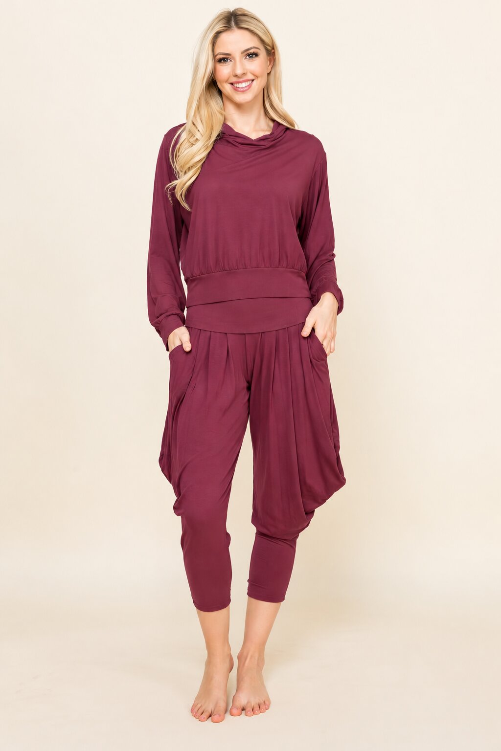 A woman modeling a matching two-piece loungewear set in a deep burgundy color, featuring a long-sleeve cowl-neck top and matching draped capri pants, against a solid light beige background.