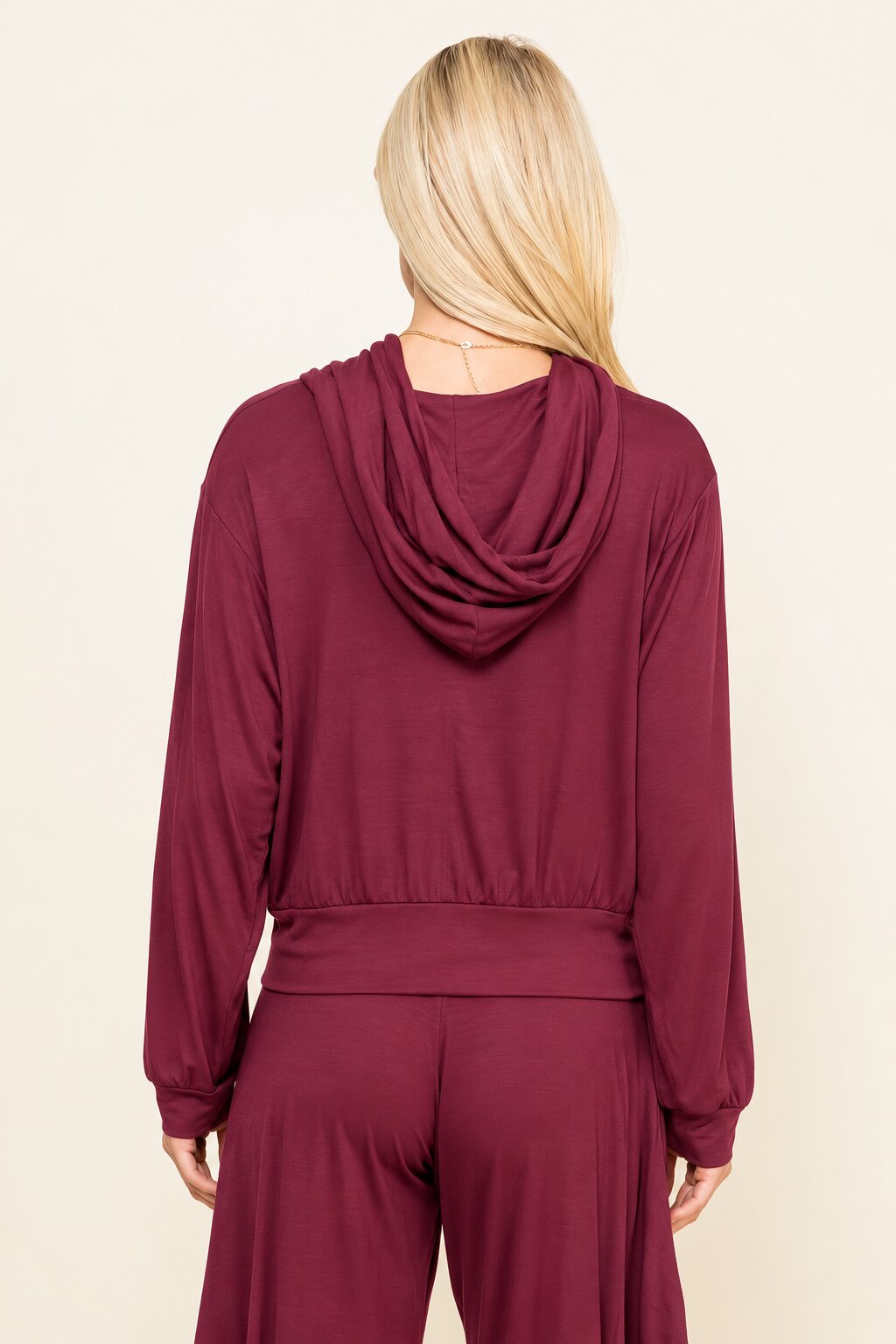 A woman viewed from the back wearing a long-sleeve draped hooded top and matching pants in a deep burgundy or maroon color, against a plain light beige studio background.