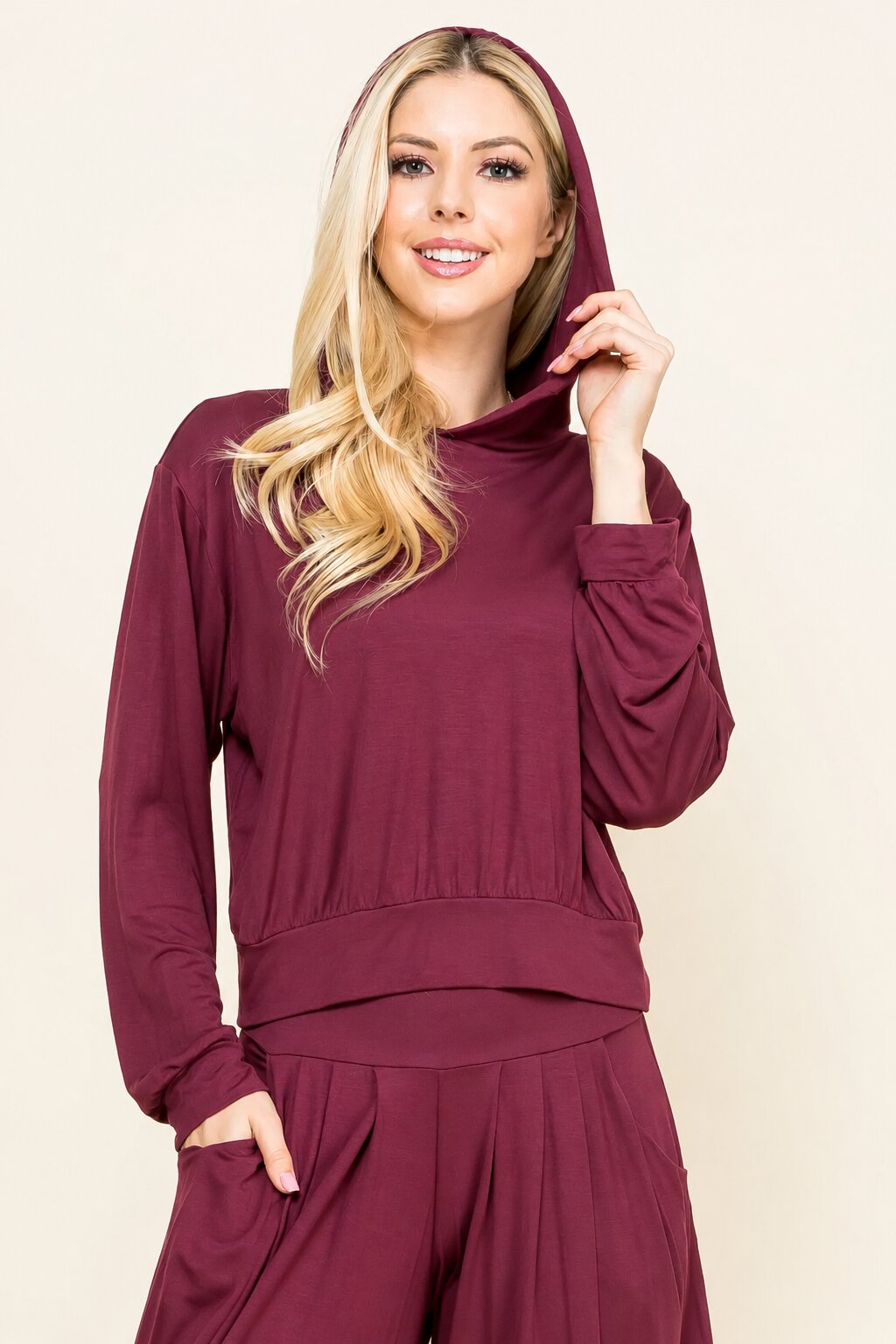 A woman modeling a matching burgundy lounge set consisting of a hooded long-sleeve top and coordinating pants, set against a plain beige background.