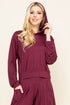 A woman modeling a matching burgundy lounge set consisting of a hooded long-sleeve top and coordinating pants, set against a plain beige background.