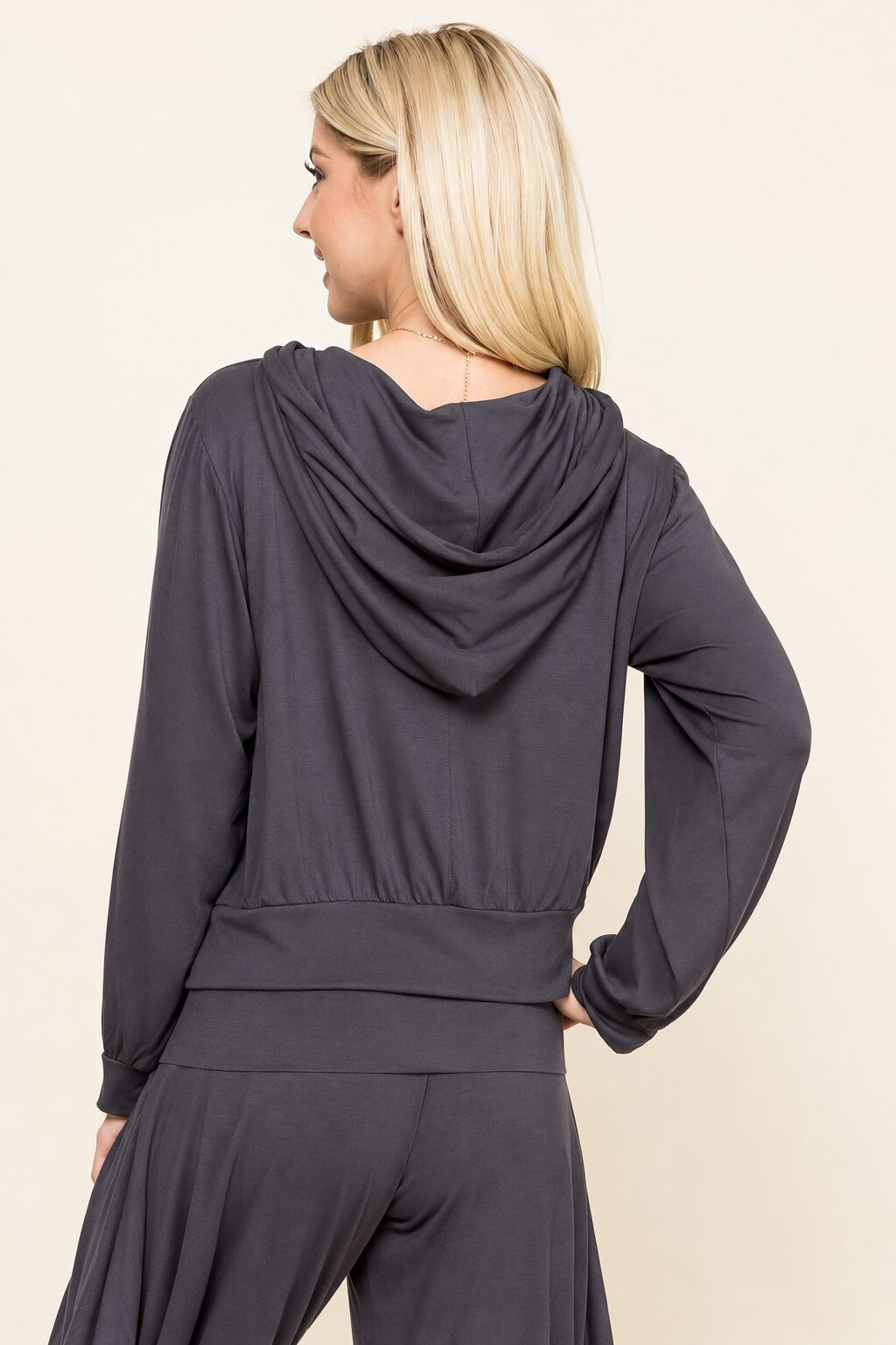 A woman shown from the back wearing a dark charcoal gray long-sleeve draped hooded top and matching pants against a plain light beige background.