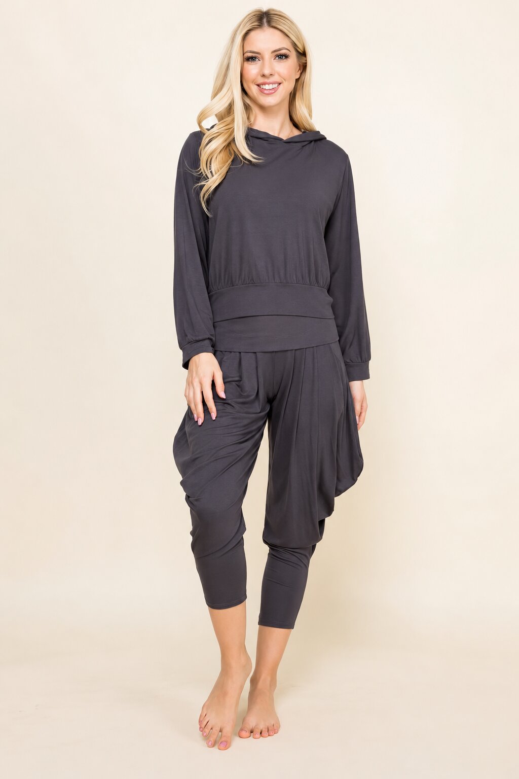 A blonde woman modeling a dark gray two-piece loungewear set consisting of a long-sleeve draped top and matching draped capri pants, standing against a solid light beige background.