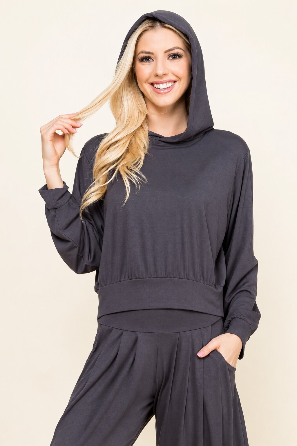 A smiling blonde woman wearing a dark charcoal gray hooded sweatshirt and matching jogger pants, posing against a solid light beige background.