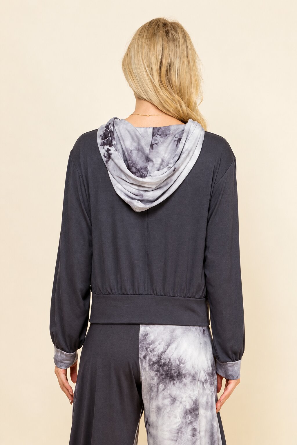 A woman shown from the back wearing a dark charcoal gray long-sleeve top with a gray and white tie-dye hood and matching tie-dye pants, against a solid beige background.