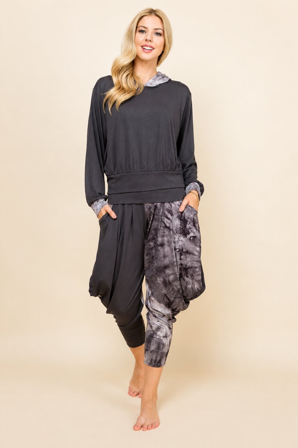 A woman modeling a dark gray lounge set consisting of a long-sleeve hooded sweatshirt with a gray patterned hood lining and matching harem-style jogger pants with a gray and black tie-dye pattern, against a solid beige background.
