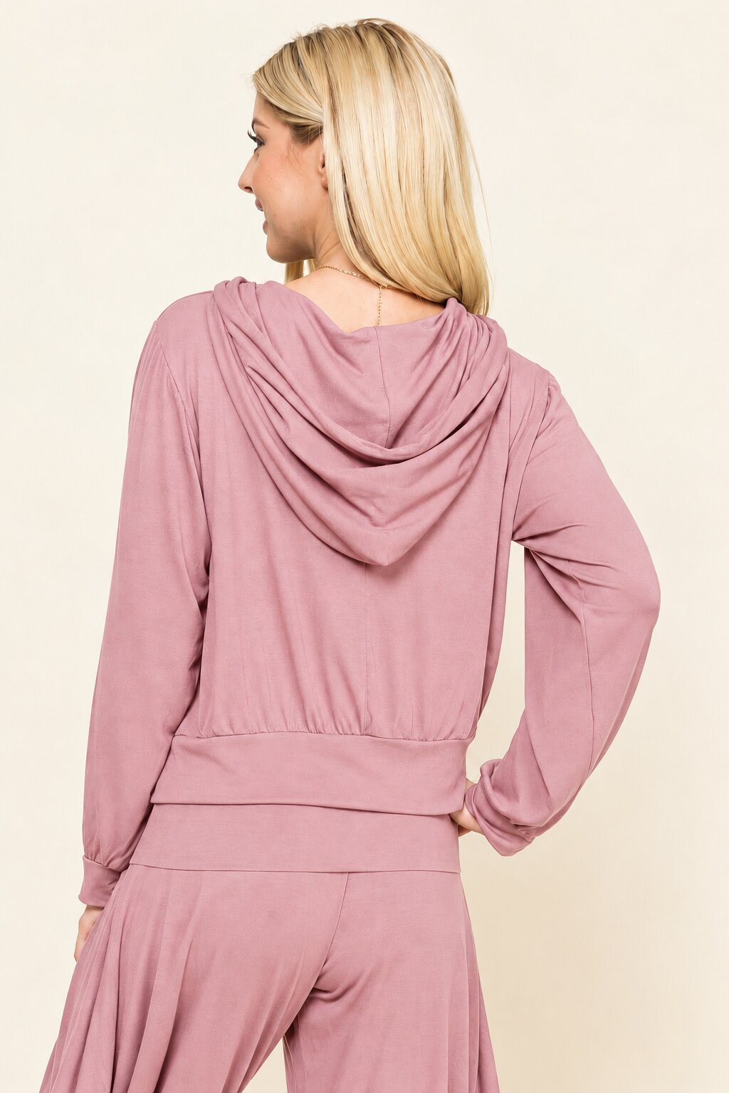 A woman shown from the back wearing a dusty rose pink long-sleeve hooded sweatshirt and matching pants against a plain cream background.