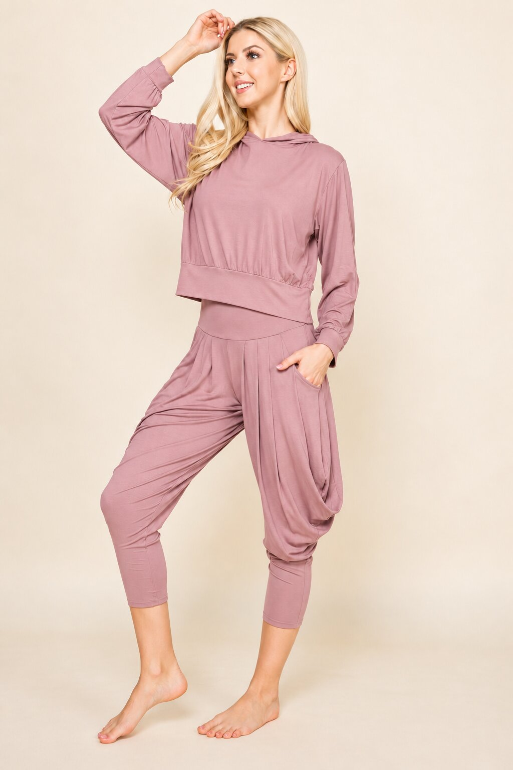 A woman modeling a dusty rose two-piece loungewear set consisting of a cropped long-sleeve hoodie and matching relaxed capri pants, posed against a solid beige studio background.