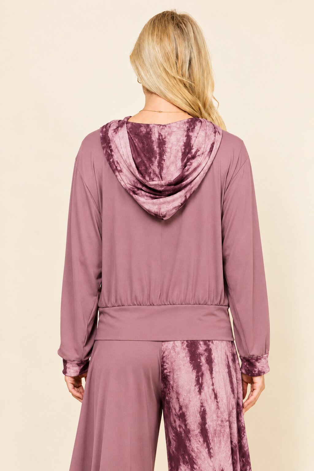 A woman shown from the back wearing a mauve-colored hooded jumpsuit with a tie-dye pattern on the hood and lower leg portion, set against a plain beige background.