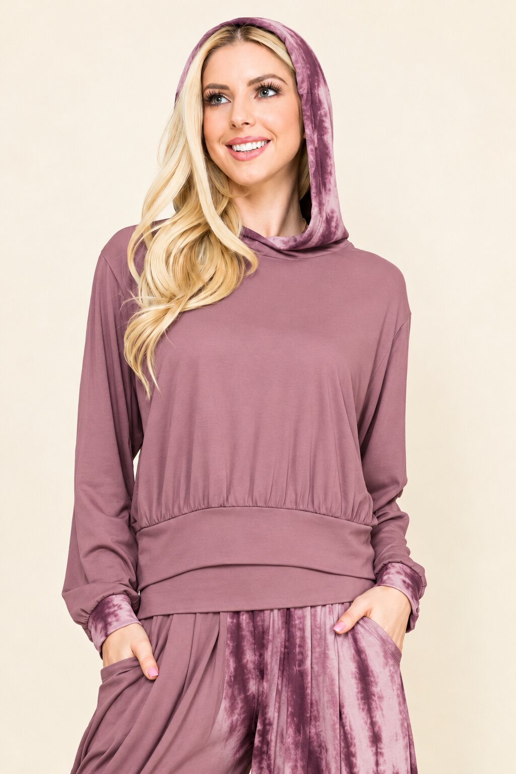 A woman modeling a matching mauve-purple loungewear set featuring a long-sleeve hooded sweatshirt and matching drawstring pants, set against a solid beige background.