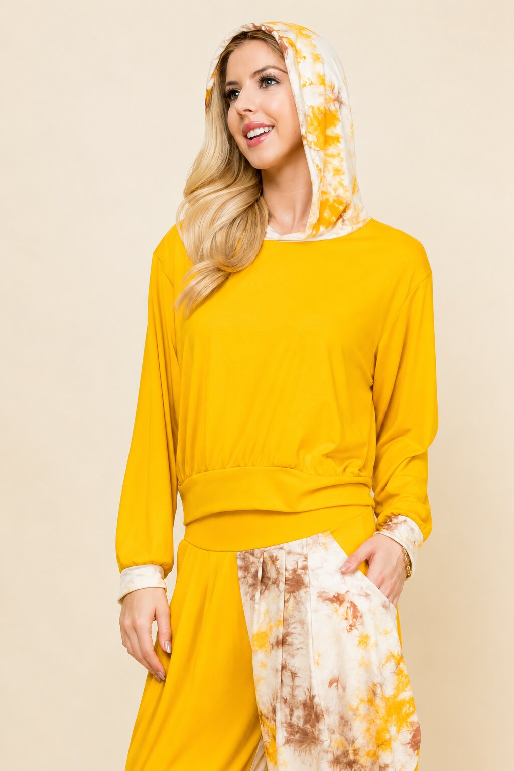 A woman modeling a bright yellow long-sleeve lounge set with a matching yellow and white tie-dye hooded sweatshirt and pants, set against a solid beige background.