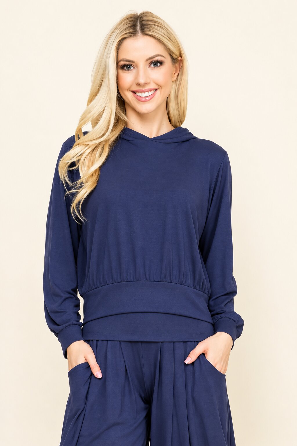 A blonde woman wearing a navy blue long-sleeve hooded sweatshirt and matching navy pants, standing against a plain beige studio background.