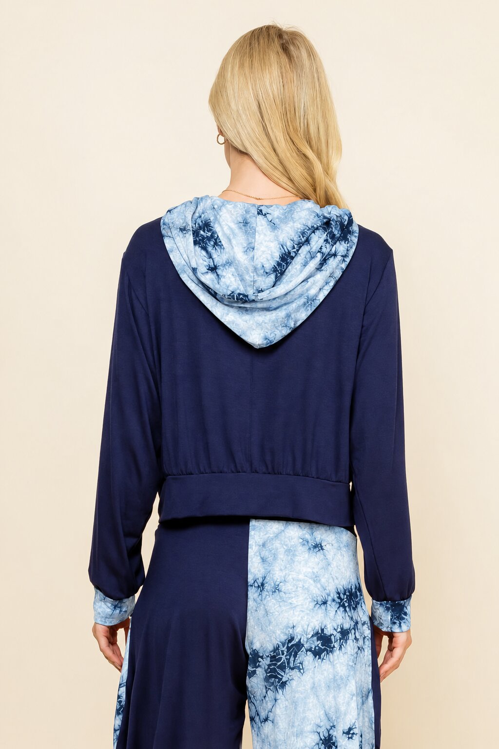 A woman shown from the back wearing a navy blue cropped hoodie with a light blue and white tie-dye hood and matching tie-dye patterned pants, against a solid beige background.
