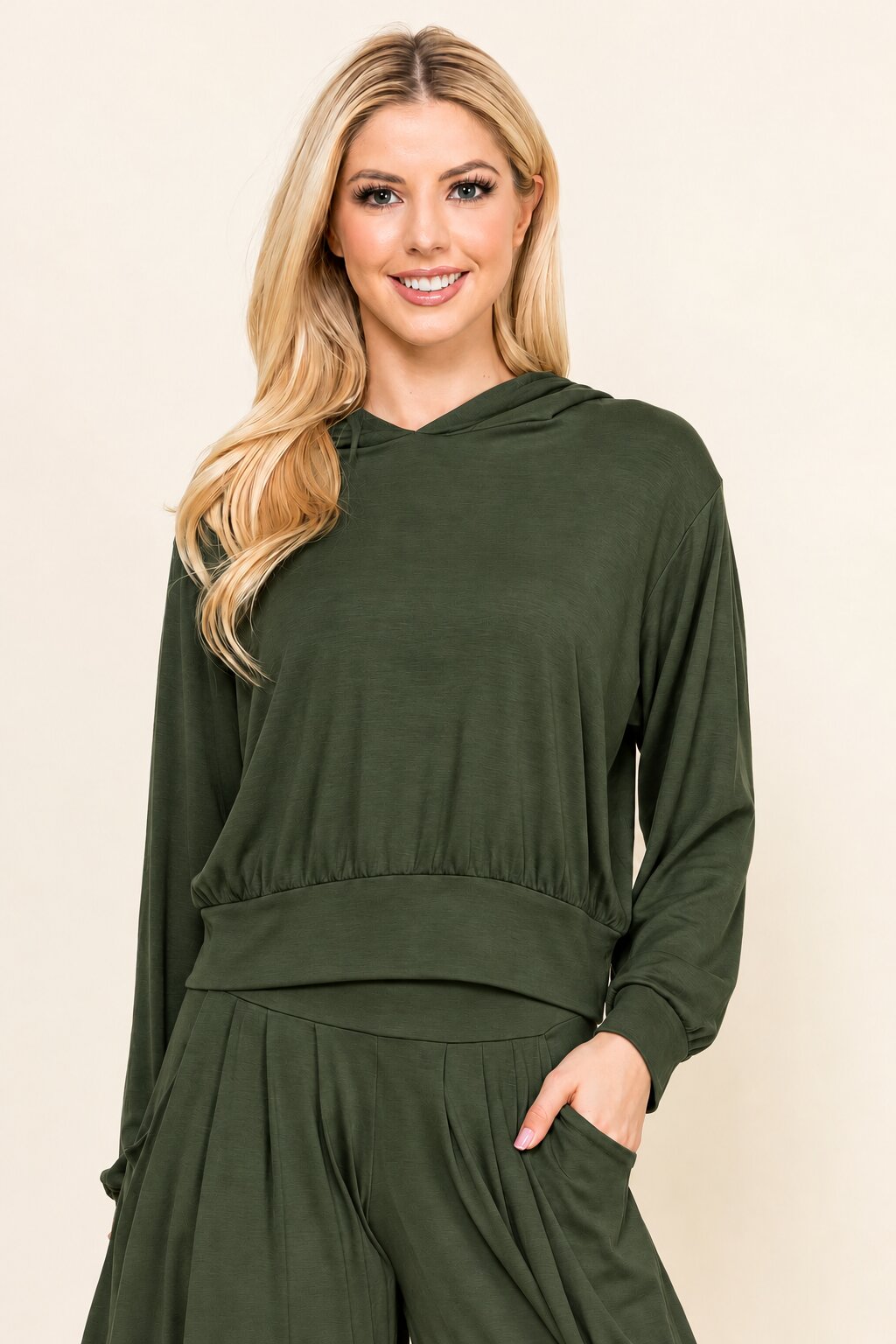 A blonde woman wearing an olive green long-sleeve hooded top and matching green pants, standing against a plain beige studio background.