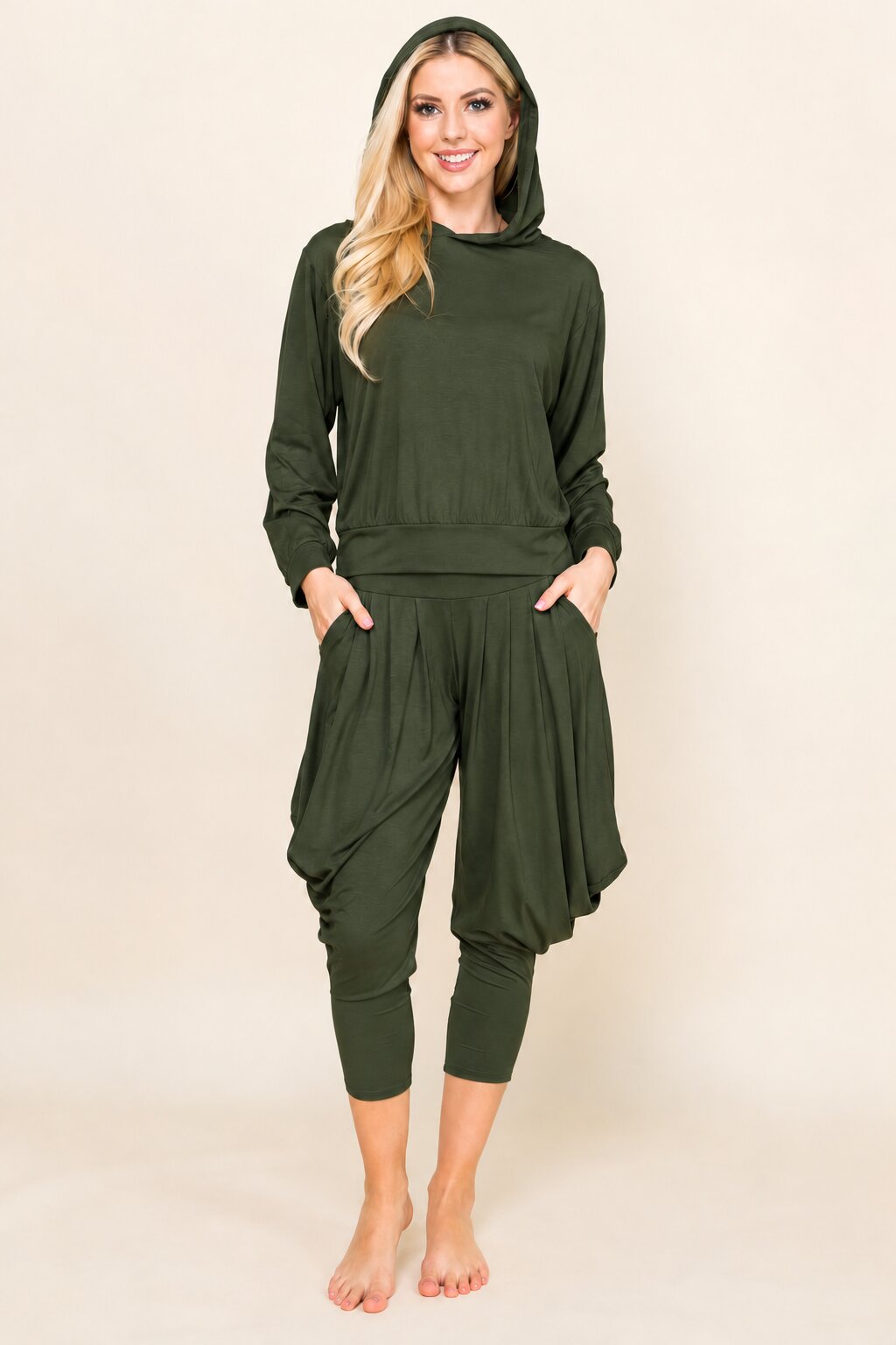A woman modeling a dark olive green hooded sweatshirt and matching baggy cropped pants with side pockets, standing against a plain beige studio background.