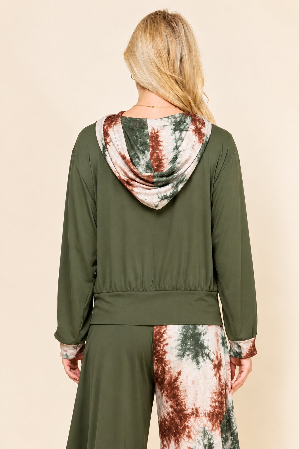 A person shown from the back wearing draped capri leggings with an olive green left leg and a brown, green, and cream tie-dye patterned right leg, against a plain beige studio background.