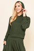 A woman modeling an olive green two-piece loungewear set consisting of a long-sleeve hooded sweatshirt and matching flared pants, set against a plain beige background.