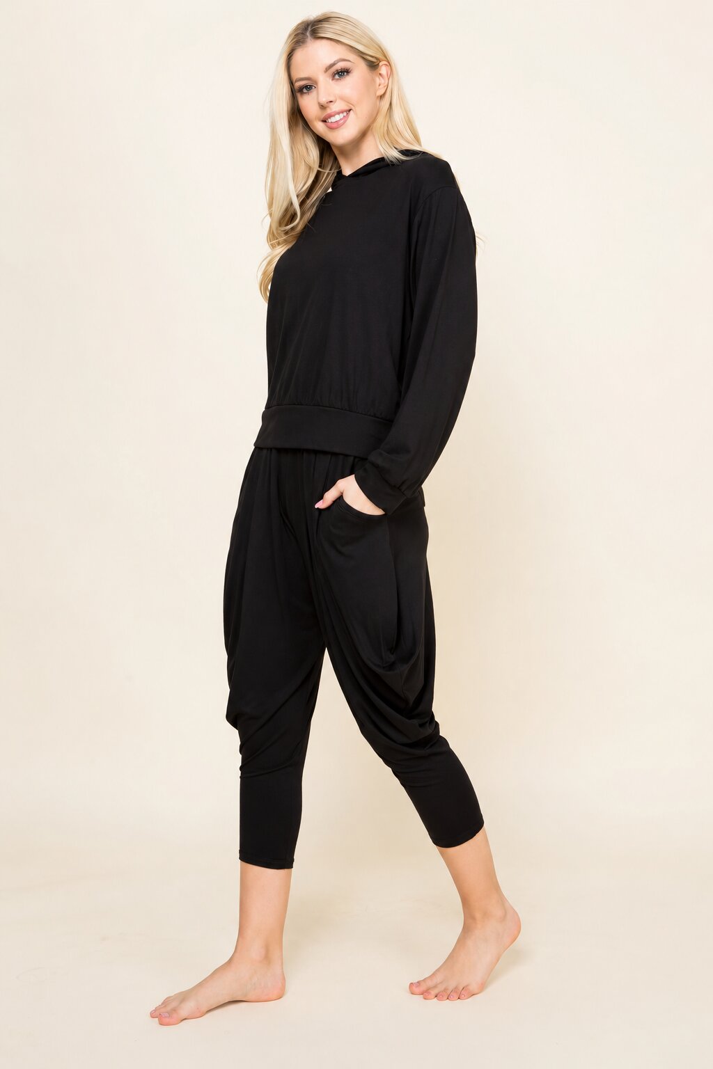 A woman modeling a black two-piece loungewear set consisting of a long-sleeve crewneck top and matching harem-style cropped pants, standing barefoot against a plain beige studio background.