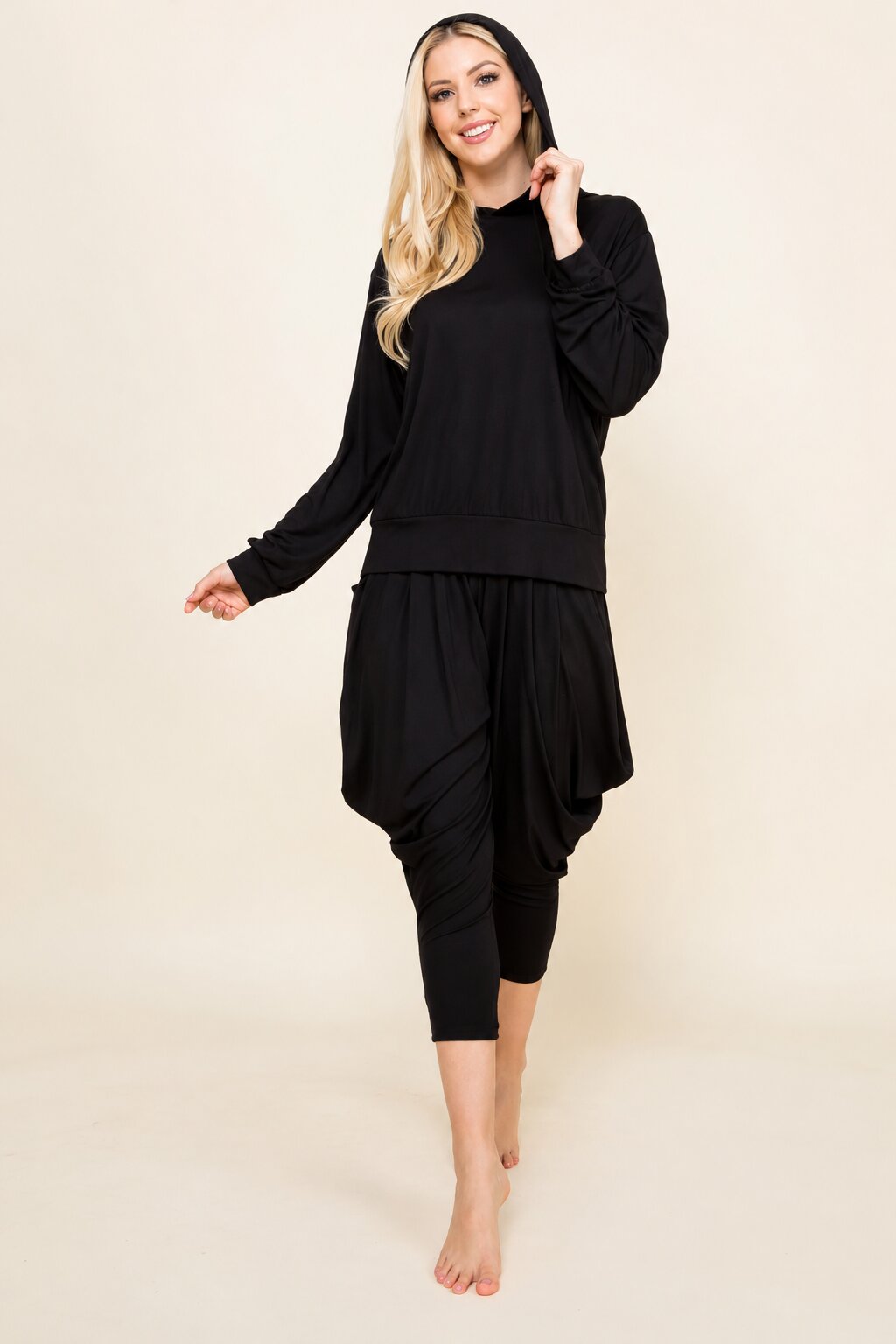 A woman modeling a black two-piece loungewear set consisting of a long-sleeve sweatshirt and matching draped capri pants, posed against a solid light beige background.
