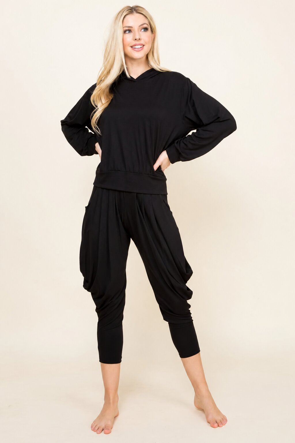 A blonde woman modeling a black two-piece loungewear set consisting of a long-sleeve cropped hoodie and matching baggy capri pants, standing against a solid beige studio background.