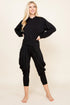 A blonde woman modeling a black two-piece loungewear set consisting of a long-sleeve cropped hoodie and matching baggy capri pants, standing against a solid beige studio background.