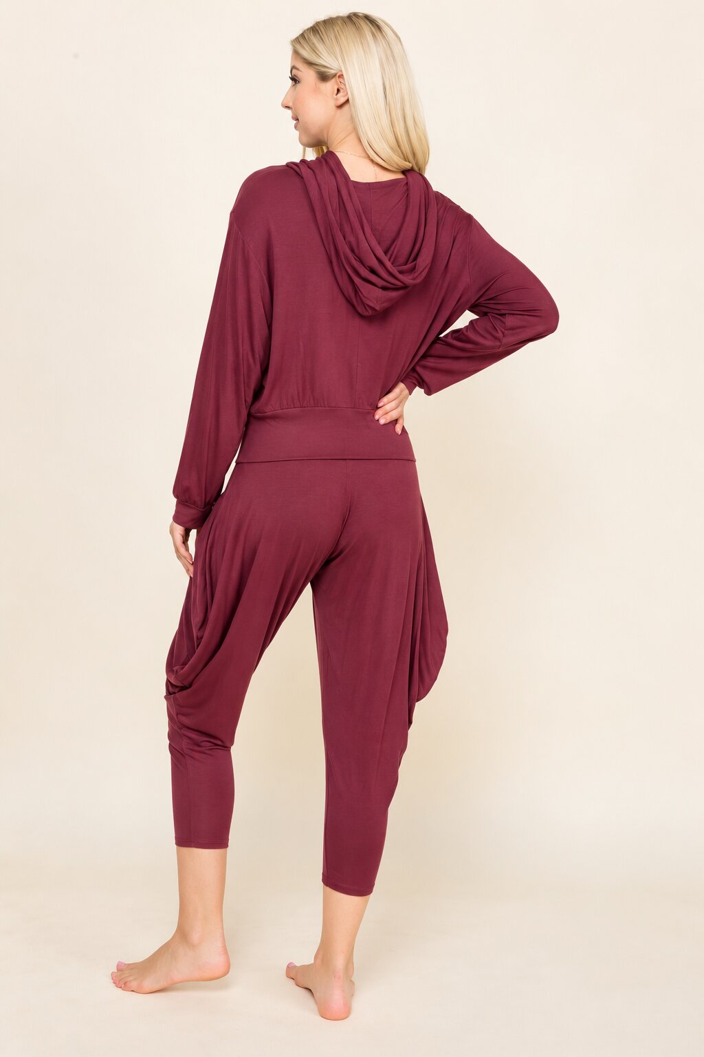 A woman modeling a matching burgundy loungewear set from the back, featuring a draped long-sleeve top and matching draped capri pants, against a solid cream background.